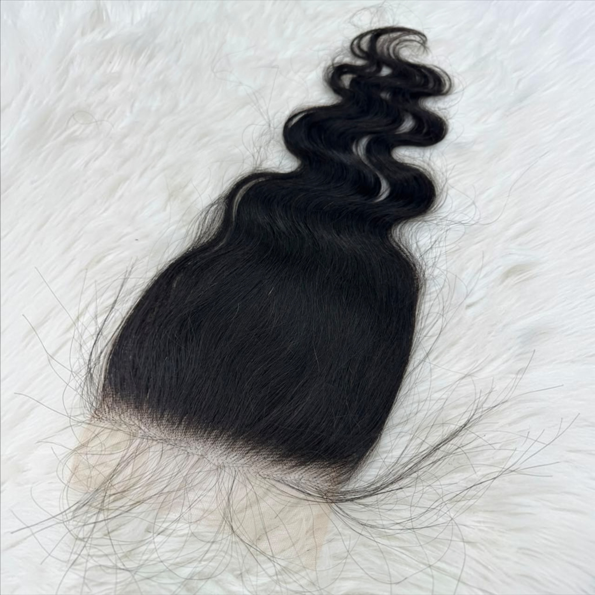 A single 5x5 HD lace closure  black body wave  with a natural-looking light beige lace base, laid flat on a white faux fur background.