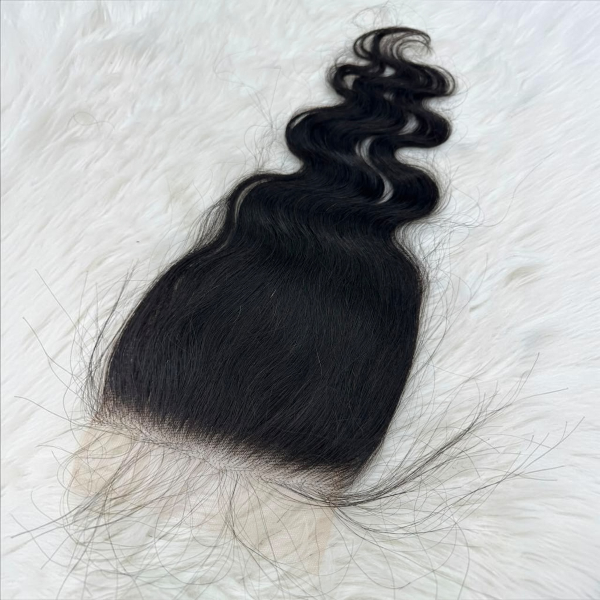 A single 5x5 HD lace closure  black body wave  with a natural-looking light beige lace base, laid flat on a white faux fur background.